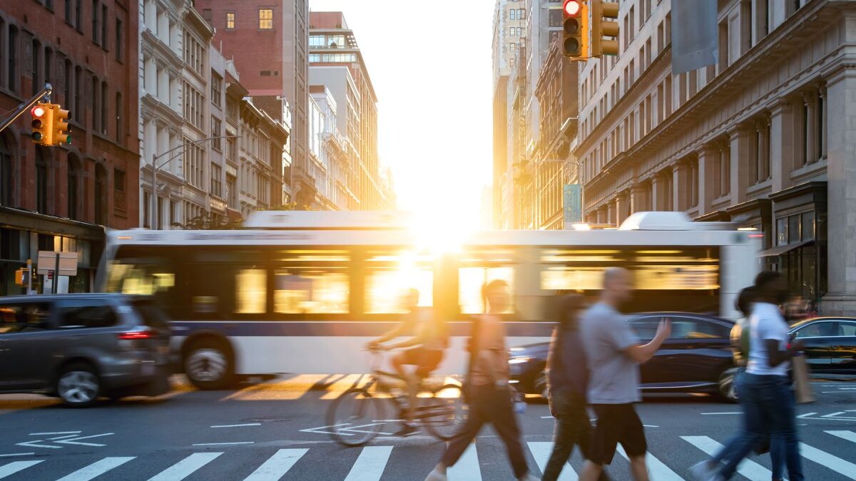 People, cars, bikes and buses traveling through a busy intersection on 5th Avenue and 23rd Street in New York City with shining sunlight background